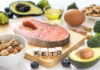A variety of healthy foods arranged on a table, including salmon, avocado, and nuts, with wooden blocks spelling 'KETO'.