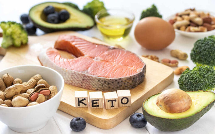 A variety of healthy foods arranged on a table, including salmon, avocado, and nuts, with wooden blocks spelling 'KETO'.