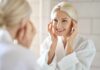 Smiling woman in a bathrobe applying skincare in front of a mirror