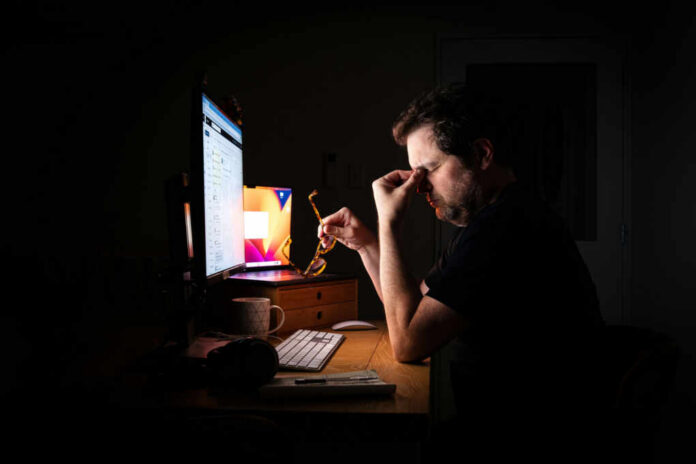 shutterstock2430605509jpg A man sitting at a desk with a computer, holding glasses and looking stressed