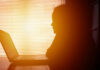 How Indoor Life Quietly Wrecks Your Body Silhouette of a person working on a laptop with sunlight streaming through blinds