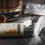 A close-up of a syringe, pills, and a glass of alcohol on a dark surface