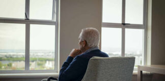 Elderly man sitting in a chair, looking out a window with a thoughtful expression