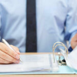 A person in a blue shirt writing on a document while holding a binder