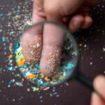 How Microplastics Hijack Our Immunities A hand holding a magnifying glass over fingers covered in colorful particles