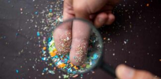 A hand holding a magnifying glass over fingers covered in colorful particles