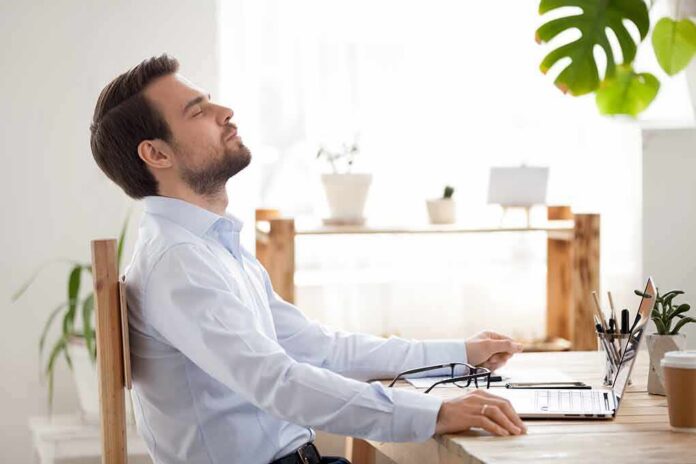 Man relaxing at desk with eyes closed beside laptop