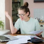 Mass Perimenopause Misdiagnosis? The Hormone Blindspot Young woman with glasses frustrated at laptop while holding bills