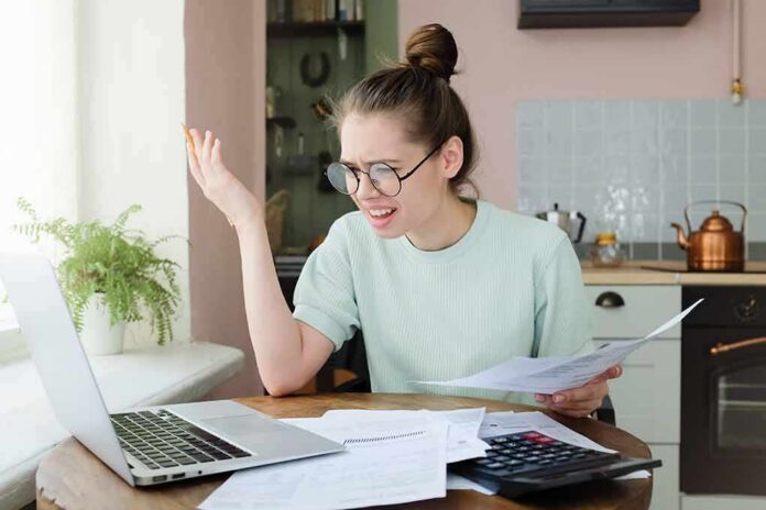 Young woman with glasses frustrated at laptop while holding bills