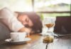 Hourglass on table with coffee cup and sleeping person in background