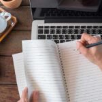 Hand writing in a spiral notebook on a laptop keyboard