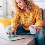 Woman on a couch using a laptop and holding a mug