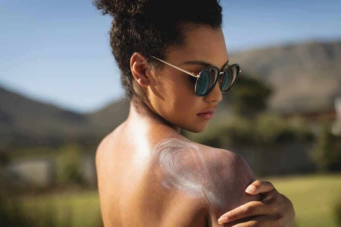 shutterstock_1313351639 Woman in sunglasses applying sunscreen to her shoulder outdoors