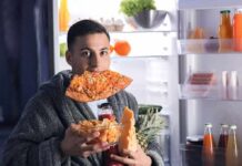 man in robe eating pizza at open refrigerator