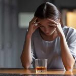 Woman with head in hands at a bar with a glass of whiskey