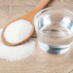 Glass of water beside wooden spoon filled with granulated sugar